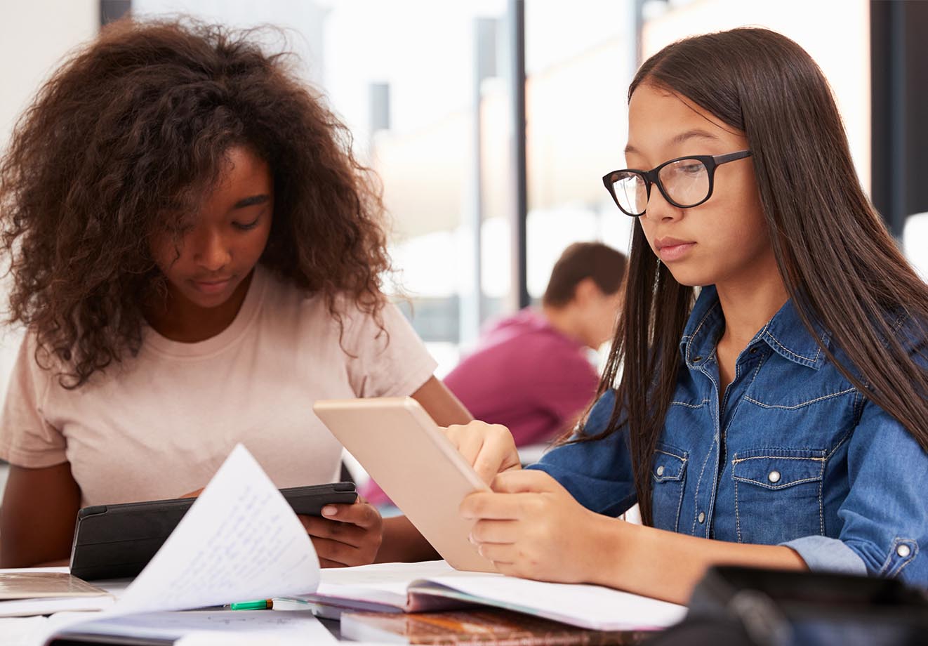 Two students working together at a table with a tablet and some books