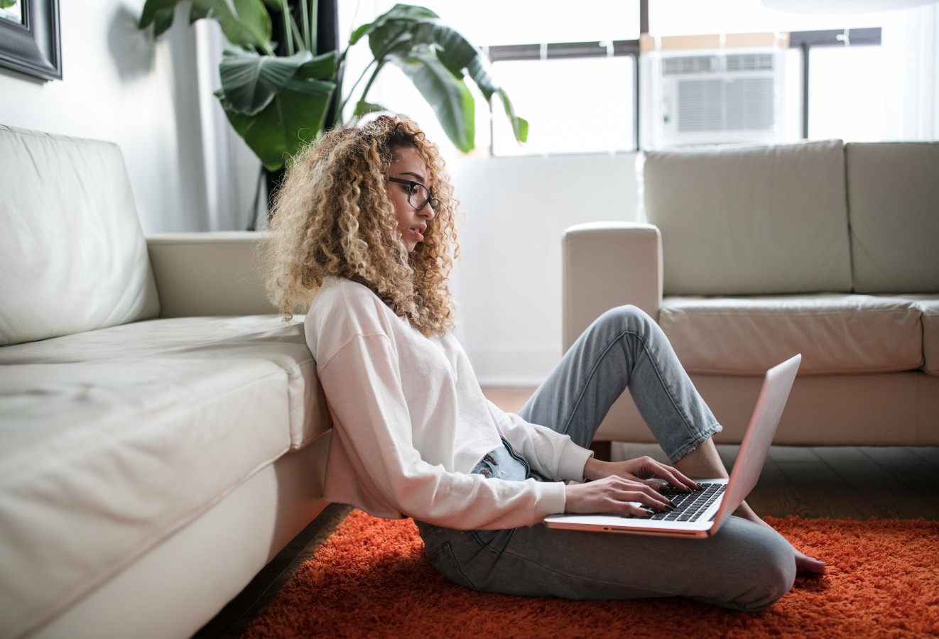 Young woman sitting on the floor, leaning against a sofa with a computer on her lap