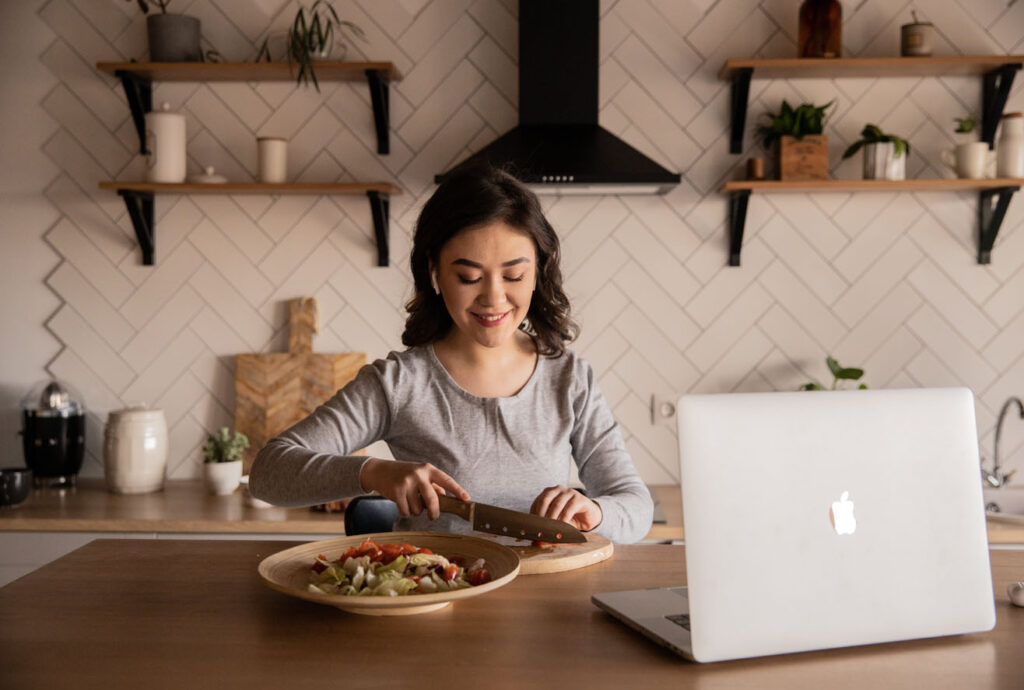 Woman chopping vegetables in kitchen as she watches laptop on counter