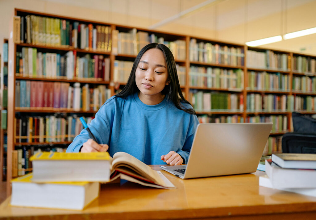 A student in a library writing notes and working on laptop