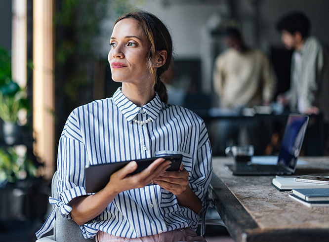 A woman holding a tablet looking thoughtfully out a window